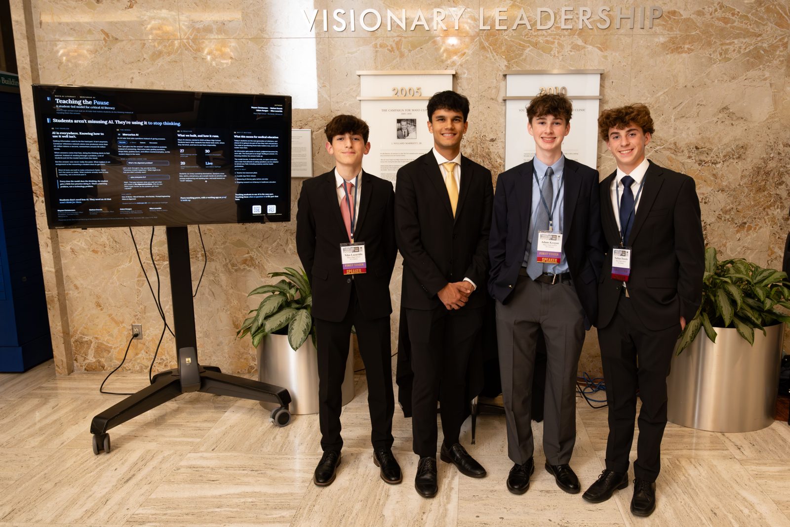 Shyam Christensen and three fellow Mayo High School students in suits standing beside a screen displaying their presentation 'Teaching the Pause' at the AACA meeting.