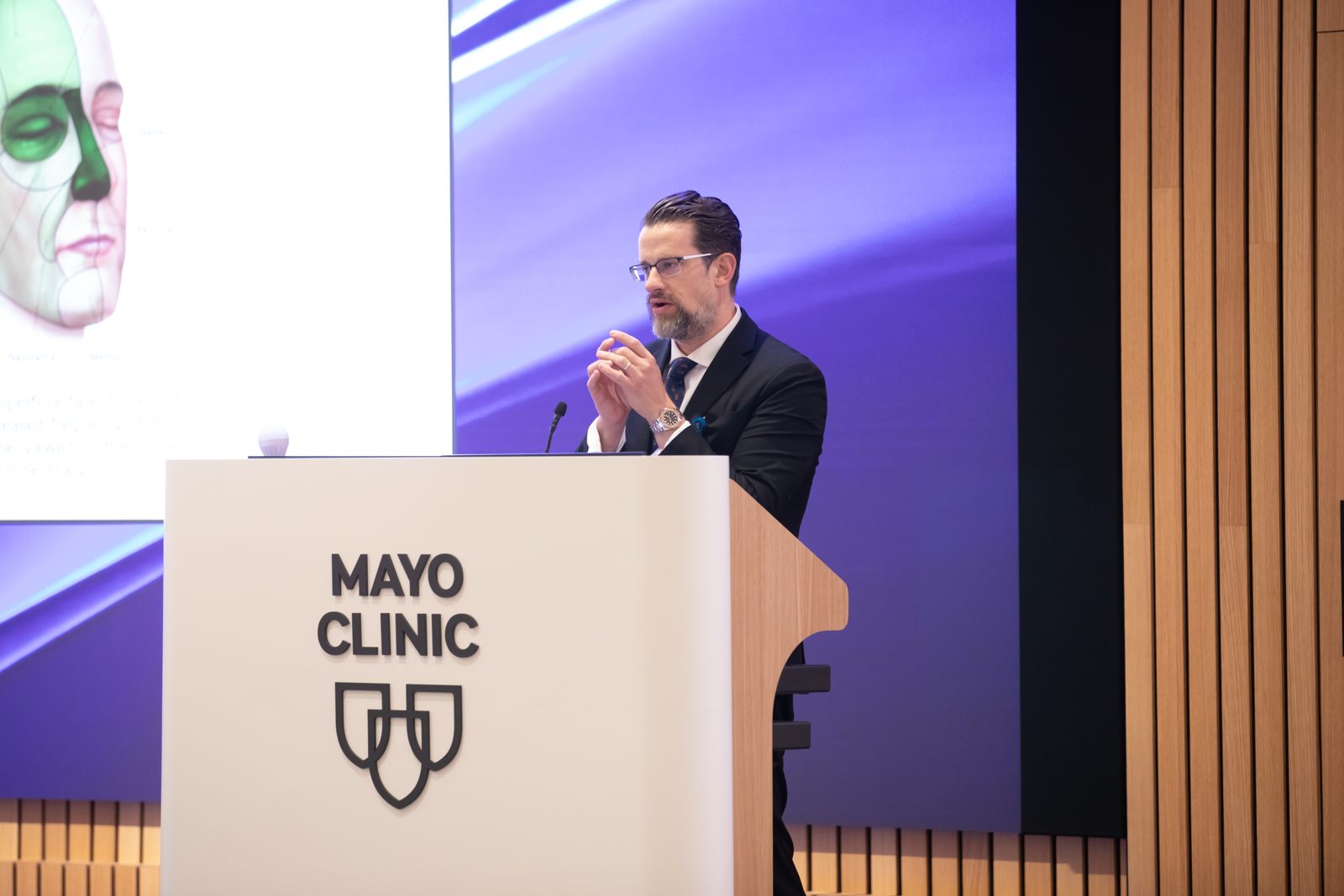 Dr. Kevin Christensen speaking at a Mayo Clinic podium during the AACA 43rd Annual Meeting, with a facial anatomy diagram on the screen behind him.