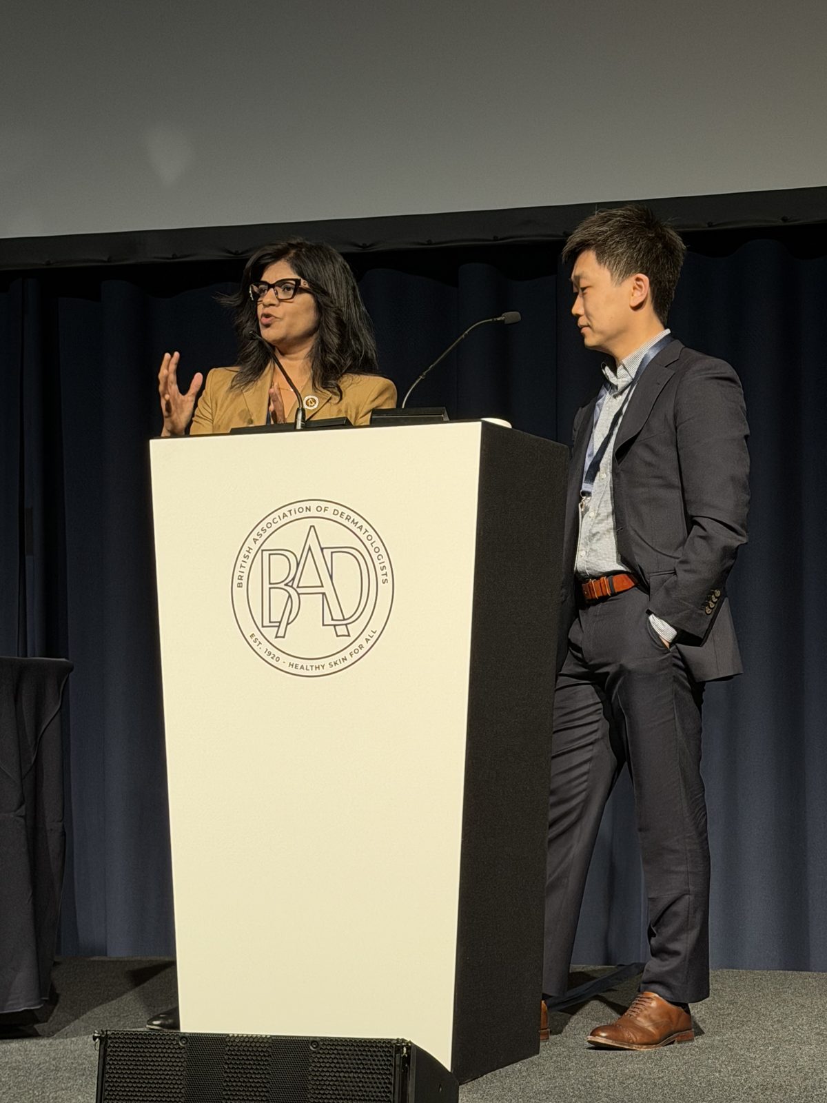 Dr. Nirusha Lachman speaking at the British Association of Dermatologists podium with Dr. Jun Zhang standing beside her.