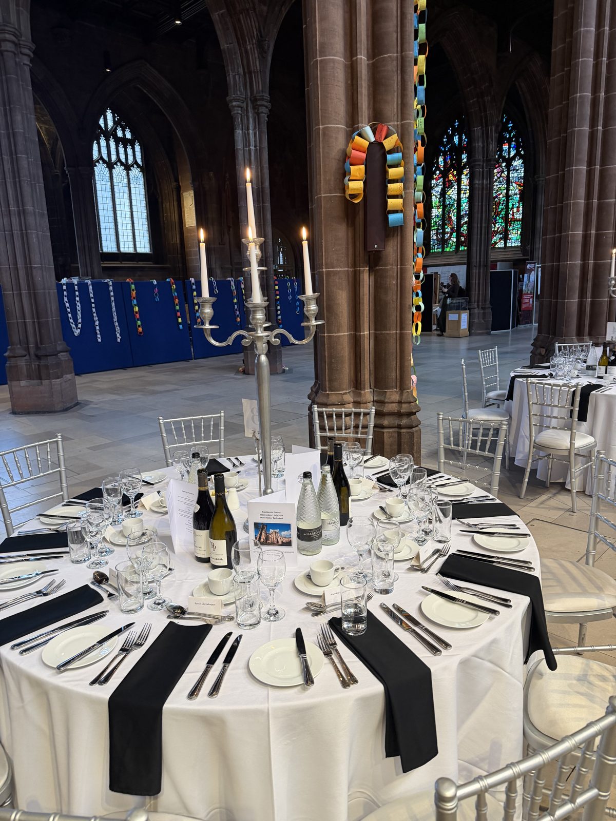 A formal dinner table with a tall silver candelabra set in the nave of Manchester Cathedral, with stained glass windows in the background.
