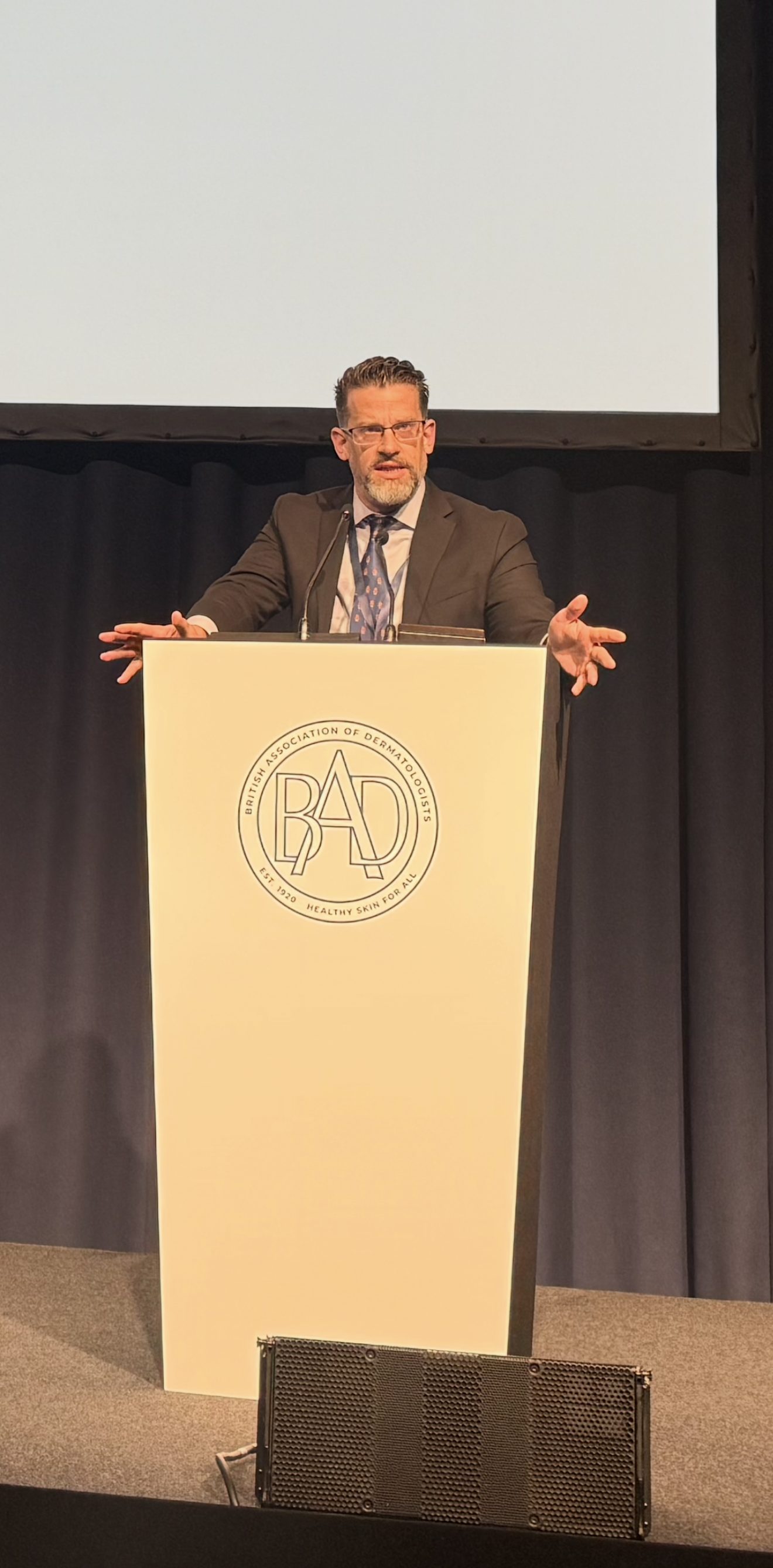 Dr. Kevin Christensen speaking with both hands outstretched at a podium bearing the British Association of Dermatologists seal.