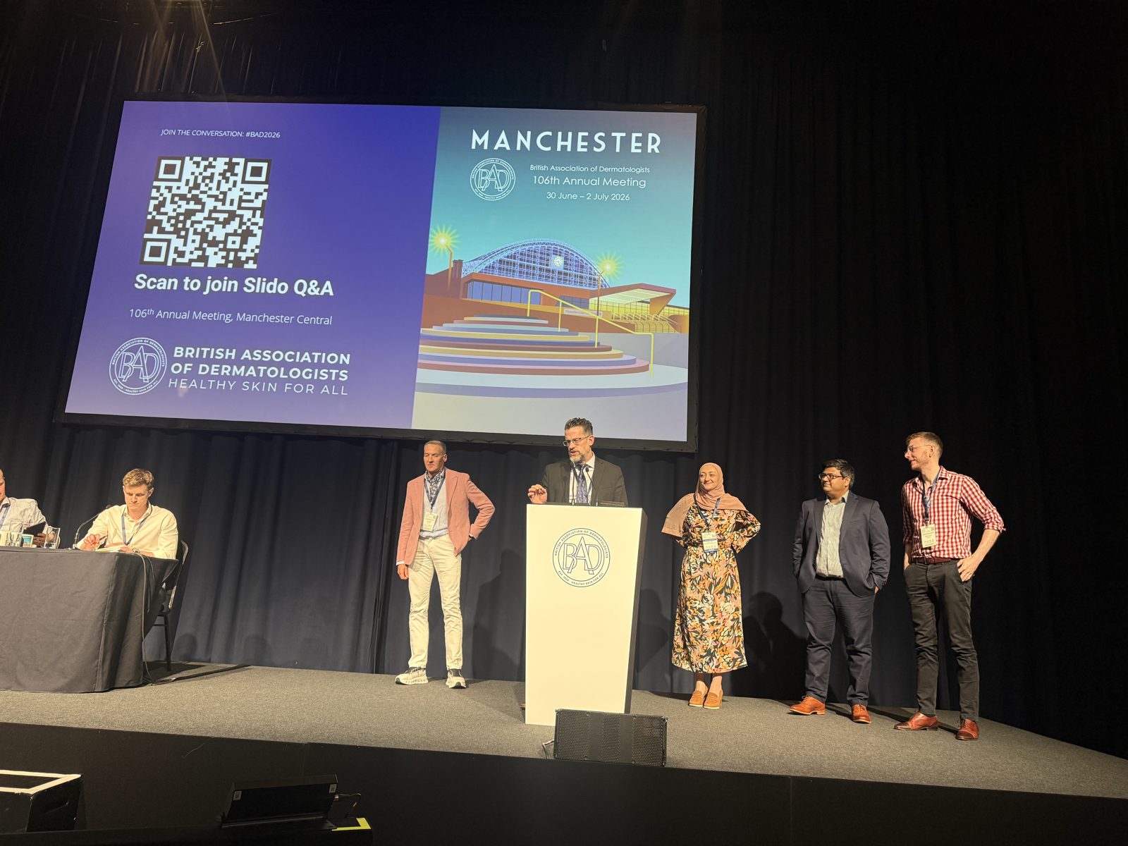 Dr. Kevin Christensen at the podium on stage during the British Society for Dermatological Surgery session, with the Manchester 106th Annual Meeting graphic on the screen and fellow speakers standing nearby.