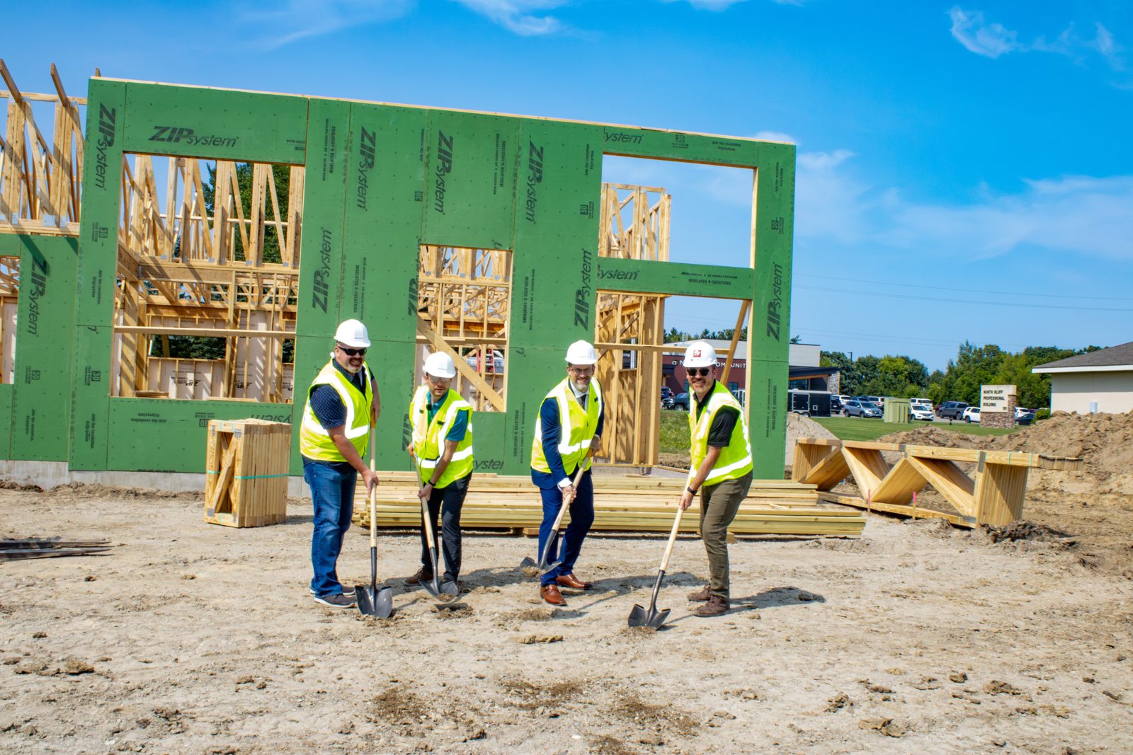 Four people in hard hats and safety vests with ceremonial shovels in front of the green-sheathed walls of the new clinic.