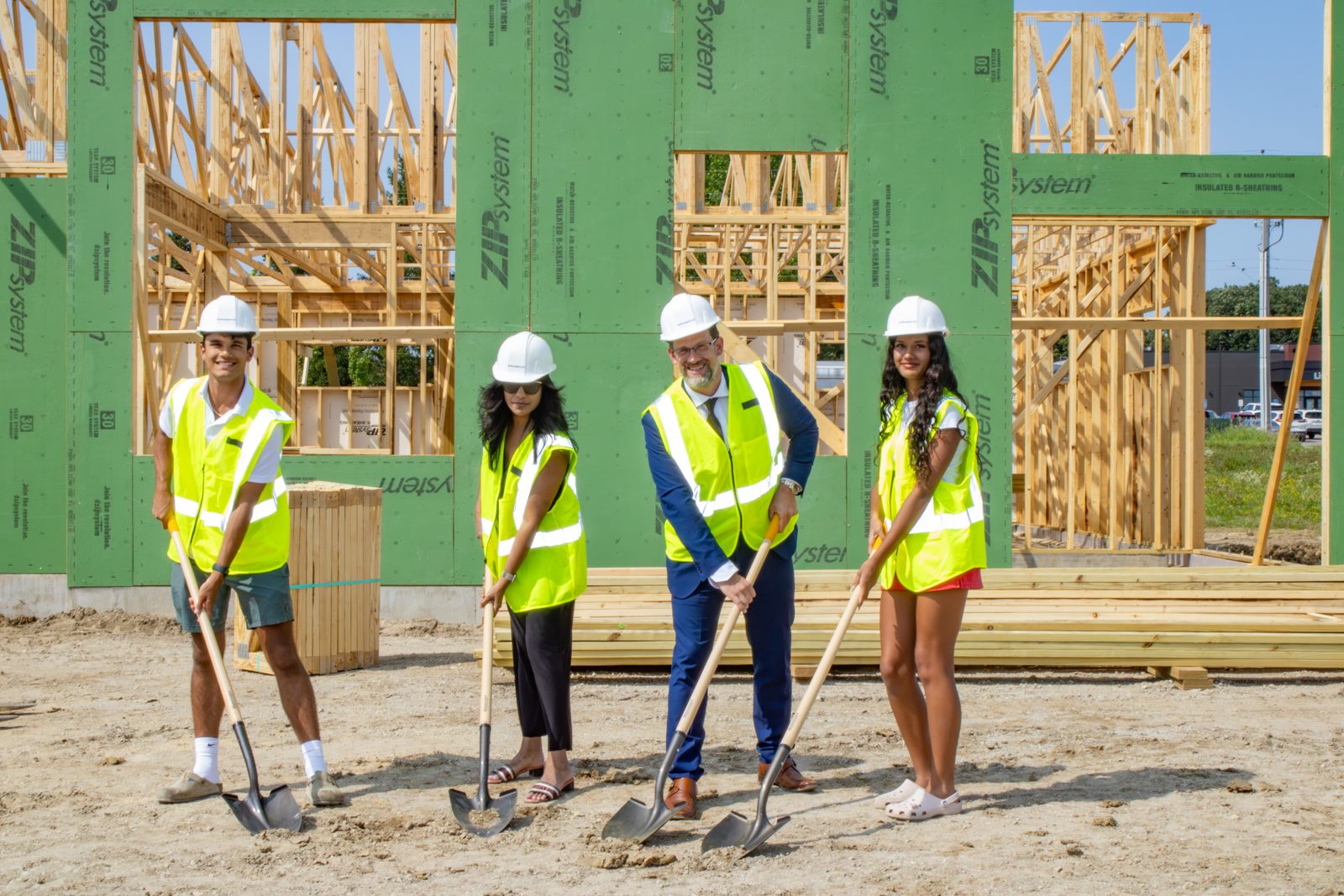 Four members of the Dermatology Partners family in hard hats and safety vests holding shovels at the Owatonna groundbreaking ceremony, with framed walls behind them.