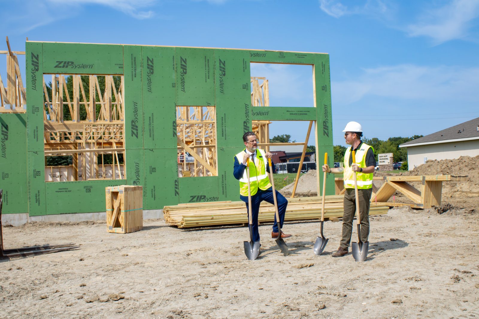 Two people in hard hats and safety vests with shovels in front of a framed wall at the construction site.